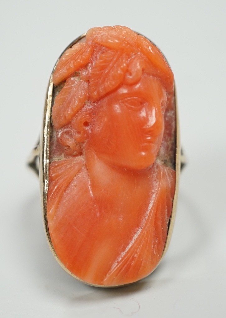 A yellow metal and coral set oval dress upfinger ring, the coral carved with a classical bust to sinister, size O, gross weight 8.2 grams.